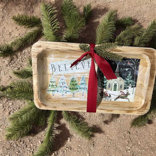 Decorative tray with Christmas scene and 'BELIEVE' text, placed on a natural surface with greenery.