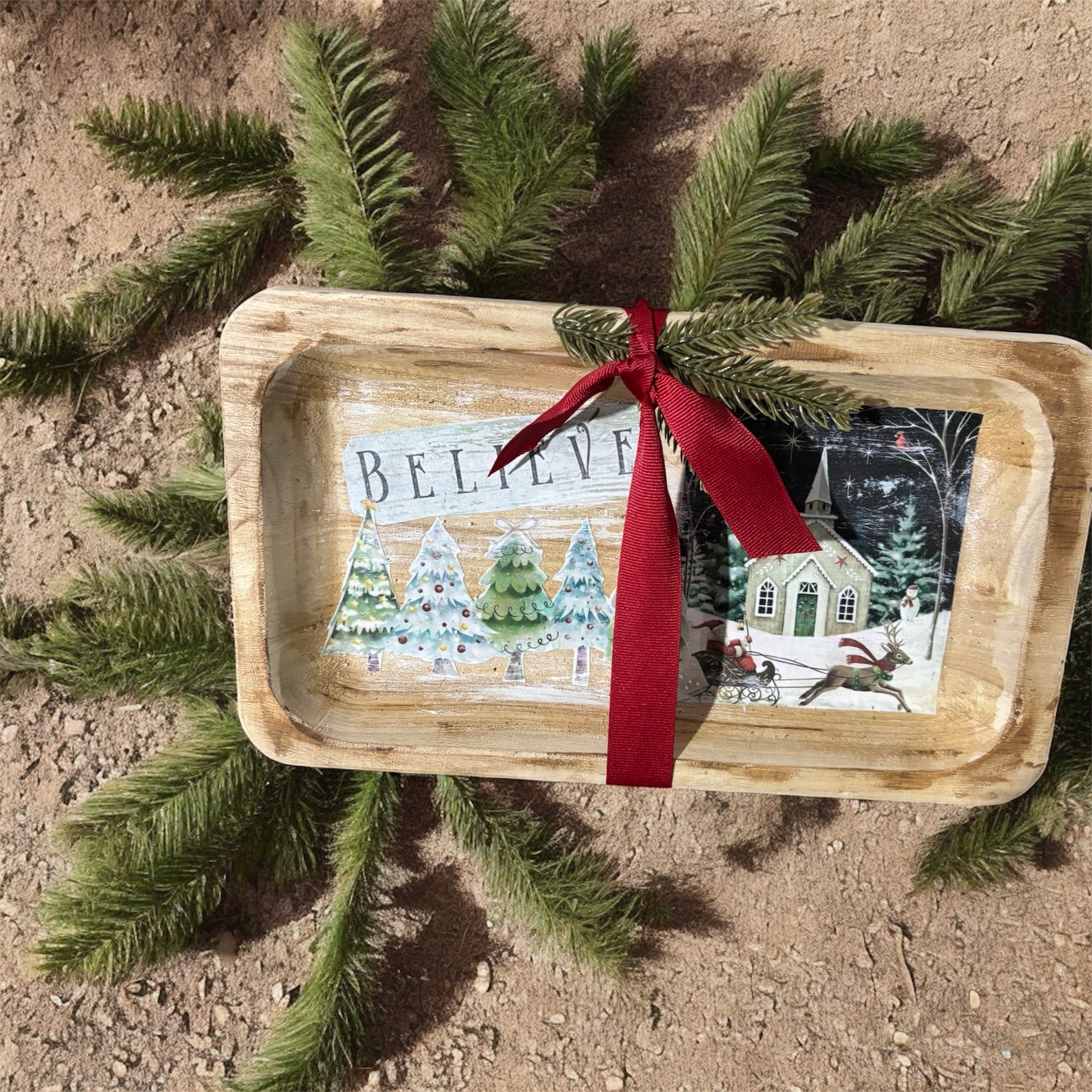 Decorative tray with Christmas scene and 'BELIEVE' text, placed on a natural surface with greenery.