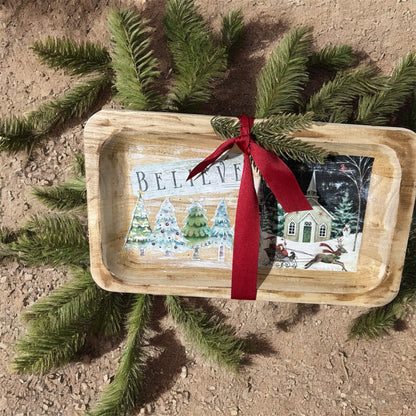 Decorative tray with Christmas scene and 'BELIEVE' text, placed on a natural surface with greenery.